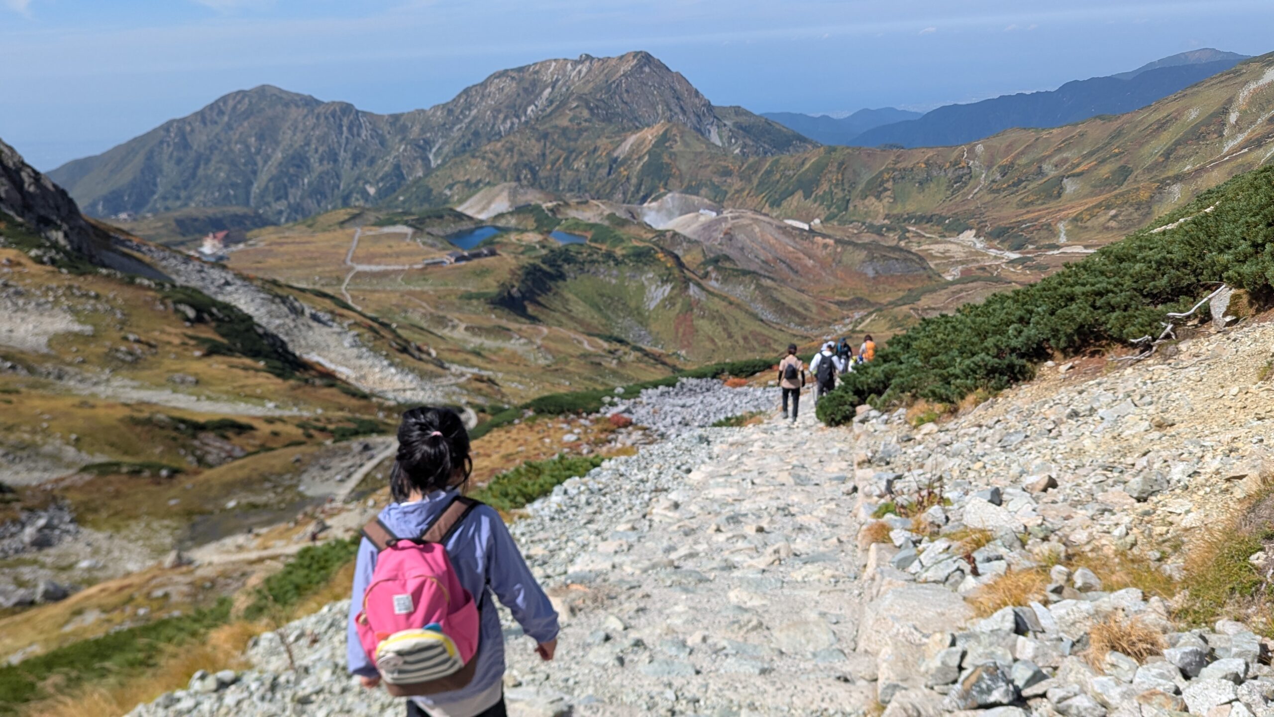 立山の下山路から室堂平を見下ろす。みくりが池と紅葉の山肌、歩く子どもの後ろ姿
