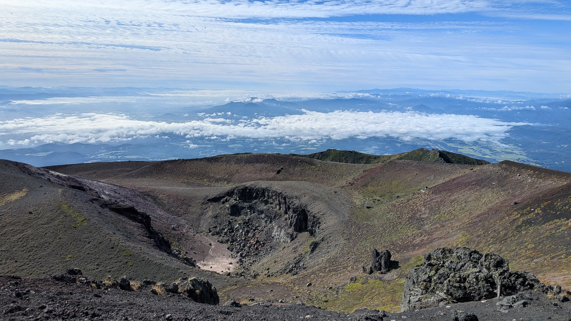 岩手山の山頂火口である御鉢を見下ろした風景。中央に小さな火口丘が立ち、奥には雲海と平野が広がる