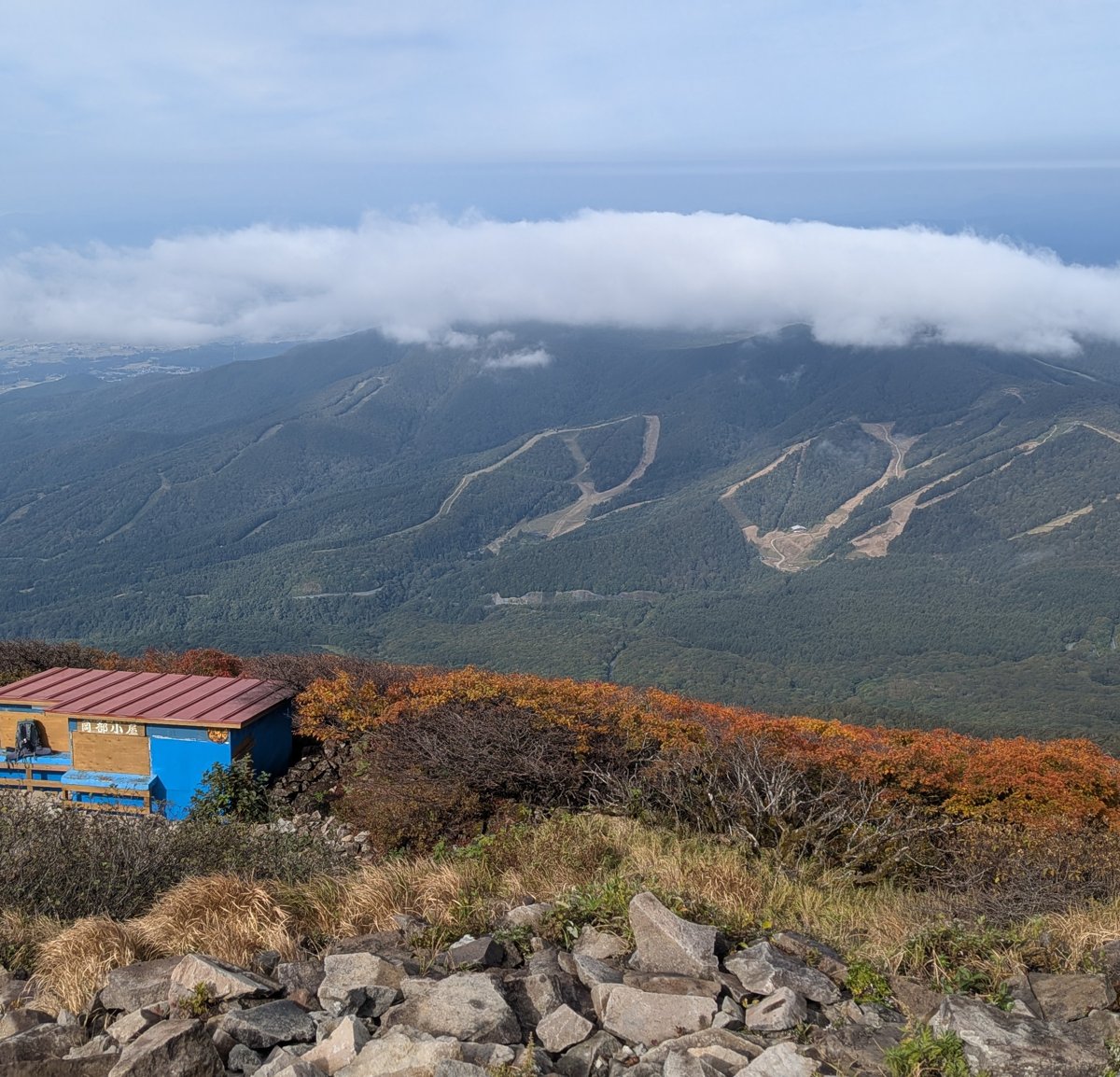 磐梯山の山頂直下にある青い岡部小屋と、西に見える猫魔ヶ岳のスキー場。手前の低木は紅葉している